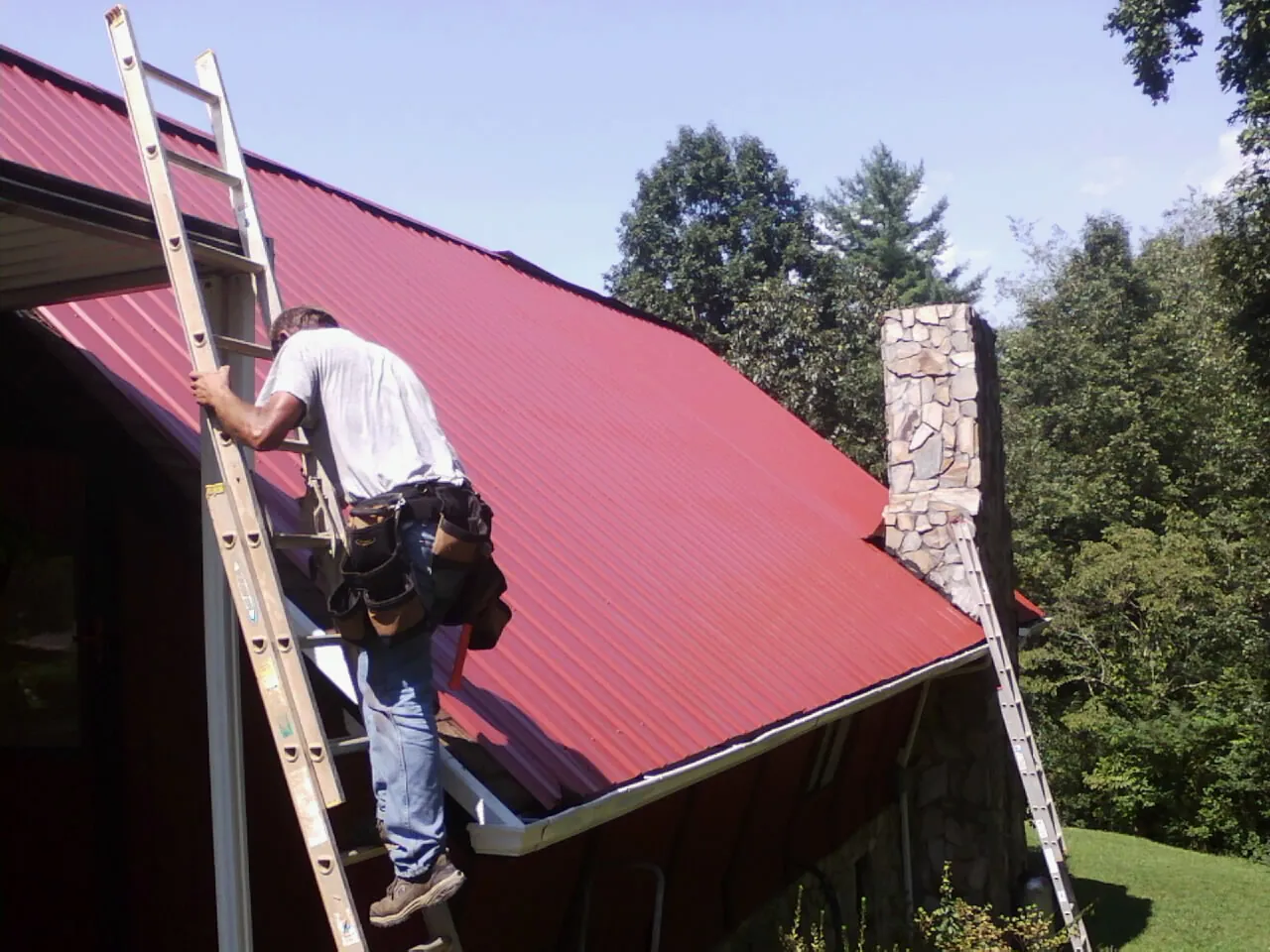 Roofer on a ladder beside a red metal roof and stone chimney — Soffit & Fascia Repair by Roofing Brothers in Garland